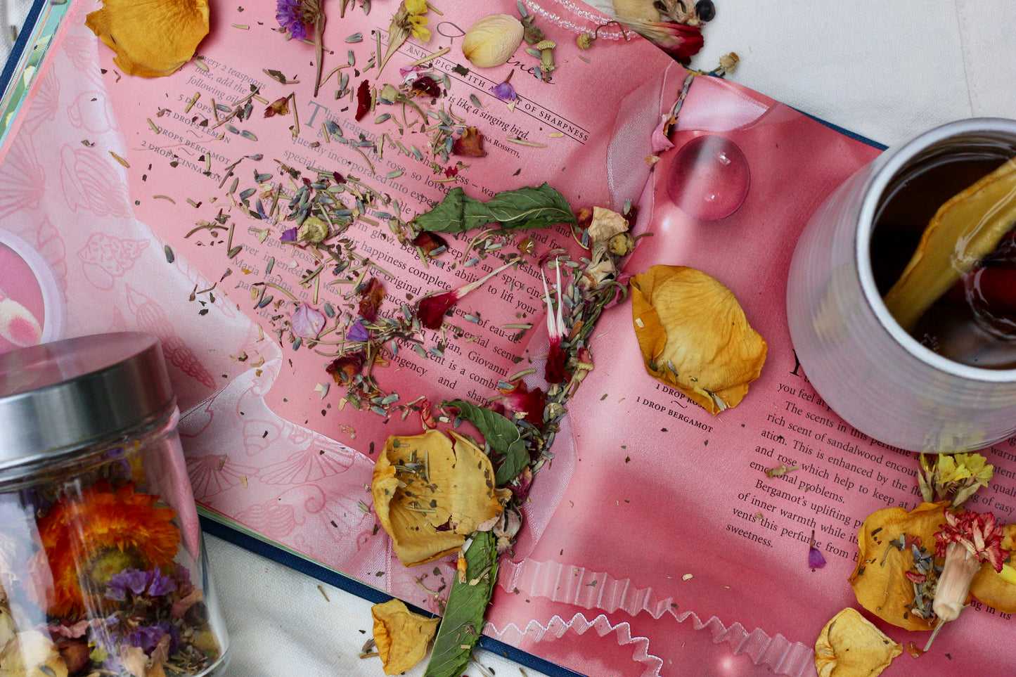 An open pink book spread with loose dried flowers and herbs scattered across the pages, alongside a small glass jar filled with colorful botanicals and a white cup containing tea and a lemon slice.
